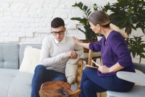 Candid shot of casually dressed professional woman psychotherapist in her fifties touching her young male patient by shoulder while having counseling session, expressing sympathy and support