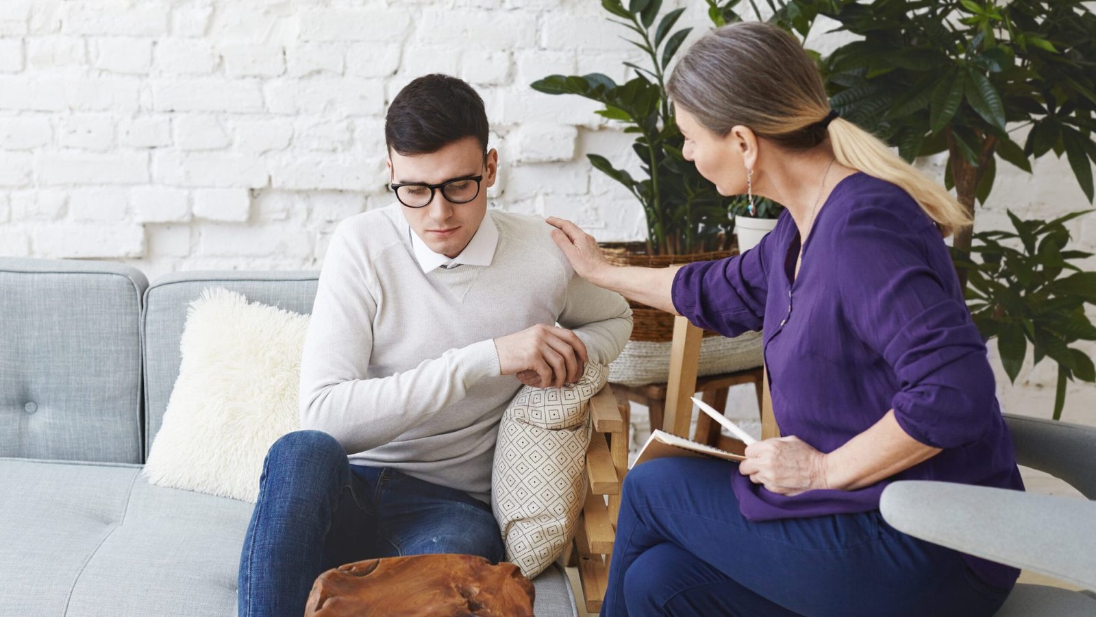 Candid shot of casually dressed professional woman psychotherapist in her fifties touching her young male patient by shoulder while having counseling session, expressing sympathy and support